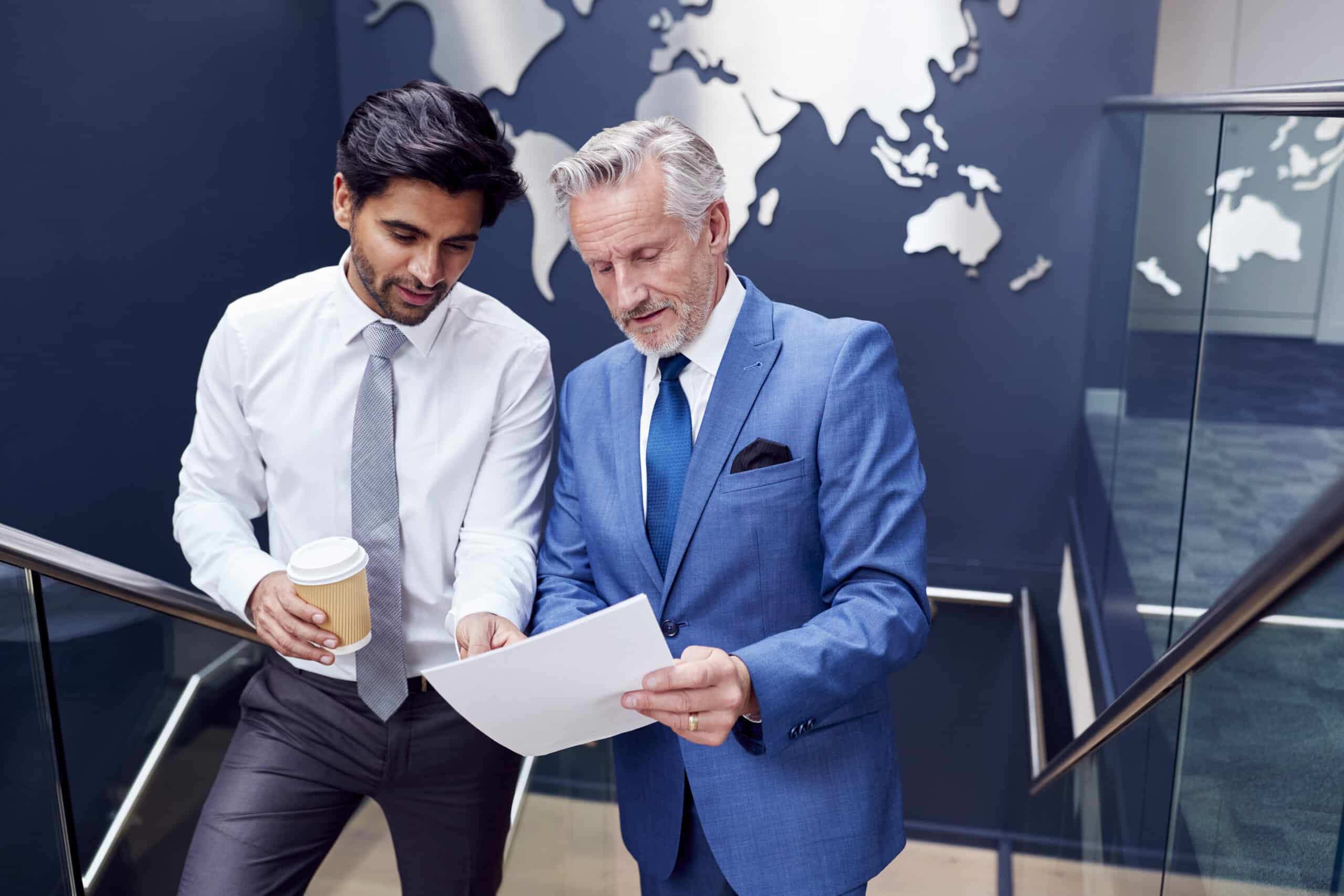 Male Colleagues Meeting On Stairs Of Office Discussing Document With World Map In Background